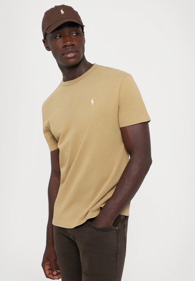 Καφέ καπέλο και μπεζ κοντομάνικη μπλούζα με μικρό λογότυπο στο στήθος, συνδυασμένα με σκούρες καφέ παντελόνα, ο μοντέλο στέκεται με τα χέρια στις τσέπες.