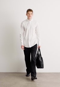 Man loopt in een minimalistische omgeving, gekleed in een witte blouse met knopen, zwarte broek, zwarte schoenen en met een zwarte leren tas.