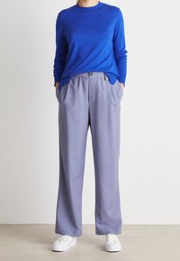 Blauer Pullover mit rundem Ausschnitt, kombiniert mit hellgrauen, weit geschnittenen Hosen mit elastischem Bund und Seitentaschen. Weiße Sneakers.