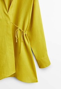 Keltainen pellavapaitatoppi, jossa on sivusolmu, pitkät hihat ja kaareva helma. Kangas on kevyttä ja sileää.