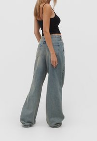 Eine Frau trägt ein schwarzes ärmelloses Cropped-Top und lockere, hellblaue Denim-Jeans und steht vor einem schlichten weißen Hintergrund.