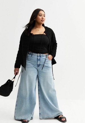 Black textured cardigan layered over a black strapless top, paired with light blue wide-leg denim jeans and a black woven handbag.