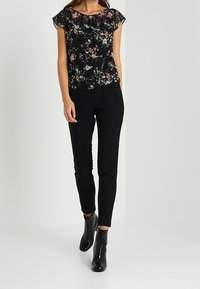 Kwiecista czarna bluzka z krótkimi, falbaniastymi rękawami, zestawiona z dopasowanymi czarnymi spodniami. Modelka ma na sobie błyszczące czarne botki za kostkę.