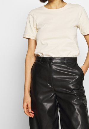 Kobieta nosząca beżową koszulę z krótkim rękawem, włożoną do czarnych błyszczących skórzanych spodni z wysokim stanem, z jedną ręką w kieszeni.