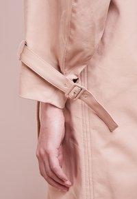 Main reposant sur le côté portant un trench-coat beige avec une sangle réglable et un détail de boucle sur la manche, sur un fond beige.