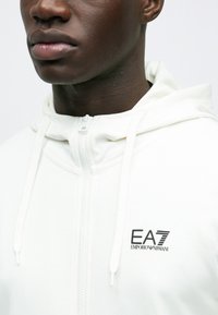 Bela jopa z zadrgo iz gladkega blaga, z vrvicami in logotipom na prsih. Prikaz bližnjice na izrez in detajle kapuce.