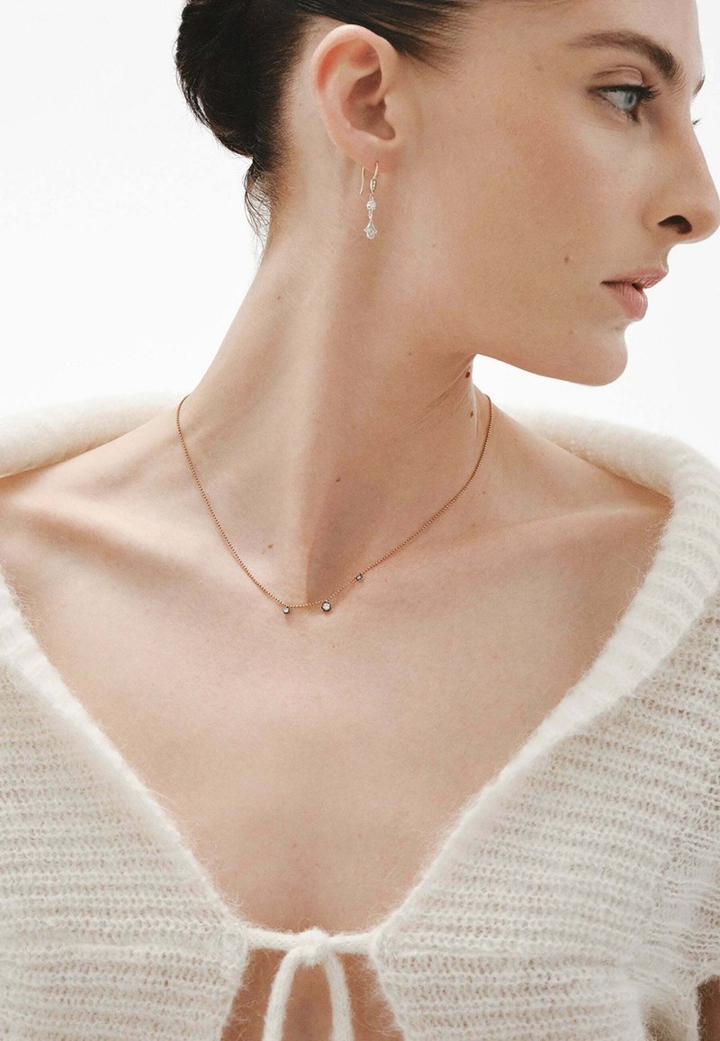 Collar de cadena de oro con pequeños colgantes redondos y en forma de lágrima, combinado con un cárdigan blanco de felpa. Los pendientes presentan un diseño similar.