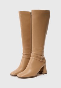 Bottes en cuir tan jusqu'au genou avec un bout carré et un talon épais. Présentent une fermeture éclair à l'avant et deux sangles décoratives avec boucles.