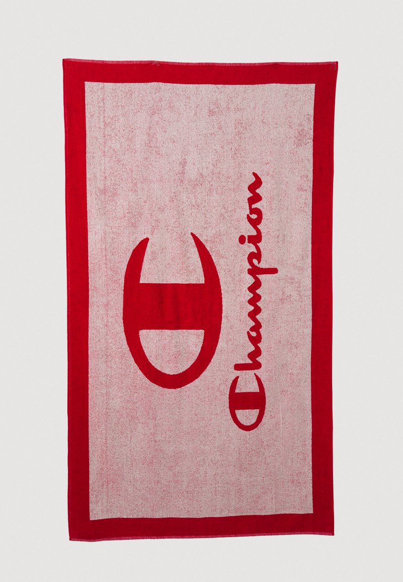 Punainen puuvillapyyhe, jossa on suuri punainen logo ja sana "Champion" rohkealla, kalligrafisella tyylillä vaaleamman punaisen taustan päällä.
