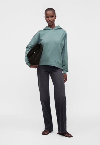 Femme debout de face portant un sweat à capuche vert, un jean foncé et des sandales, tenant un sac matelassé sombre sous le bras, sur un fond neutre.