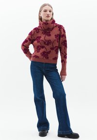Strick-Hochkragenpullover in Rost mit floralem Muster in Dunkelrosa, kombiniert mit weit geschnittenen blauen Jeans und schwarzen Stiefeletten.