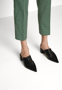 Zwarte leren muiltjes met een spitse neus en een lage blokhak. Gemusterde groene broek met witte vierkanten en een cropped lengte.