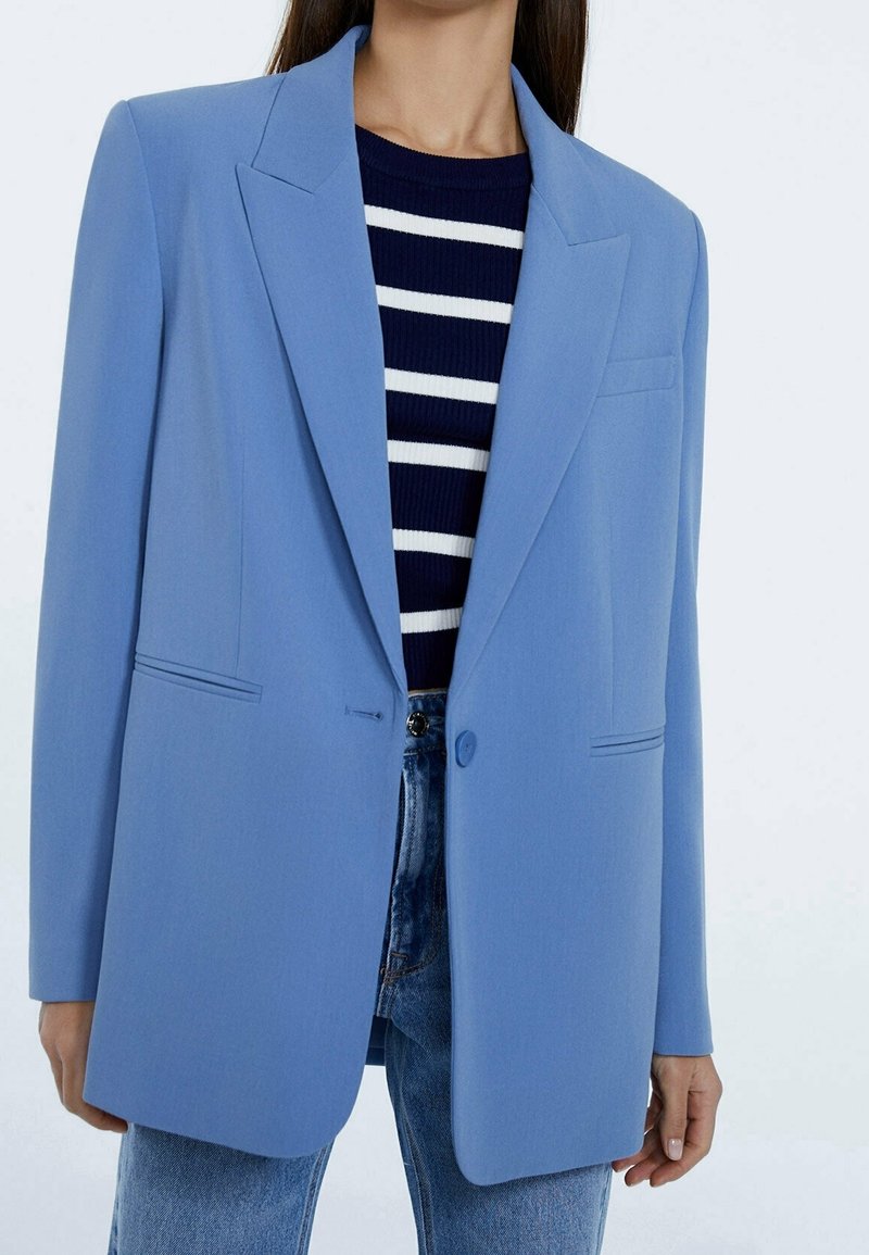 Ljust blå blazer med en strukturerad design, enkel knäppning och två sidofickor, bärs över en marinblå och vit randig tröja.