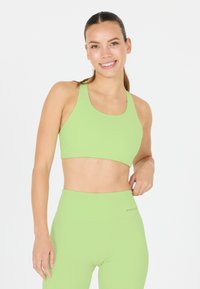 Jeune femme souriante, portant un soutien-gorge de sport vert citron et un legging taille haute, debout devant un fond blanc uni.