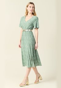 Zielona, kwiatowa sukienka midi z krótkimi rękawami i dekoltem w kształcie litery V, ściągnięta w talii metalowym paskiem. Posiada boczne rozcięcie oraz zwiewny materiał.