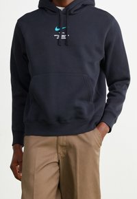 Marinblå hoodie i mjukt tyg med en känguruficka, med en framträdande turkos Nike-logotyp och grafisk text på framsidan.