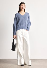 Ανοιχτό μπλε πλεκτό πουλόβερ με V-λαιμόκοψη και φαρδιά μανίκια, συνδυασμένο με άνετο λευκό παντελόνι και μικρή μαύρη και ασημί τσάντα.
