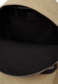 Rucsac din corduroy bej, cu o deschidere cu fermoar, căptușeală interioară neagră și un mic patch din piele cu logo în interior. Detaliu din hardware argintiu al fermoarului.