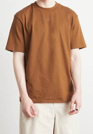 Homme portant un t-shirt HUGO marron à manches courtes et un pantalon beige clair, debout avec les bras détendus le long du corps.
