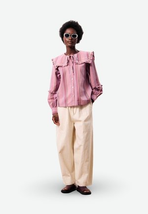 Femme portant des lunettes de soleil blanches, un chemisier rayé rose avec un col à volants et un pantalon large crème, debout avec une main dans la poche.