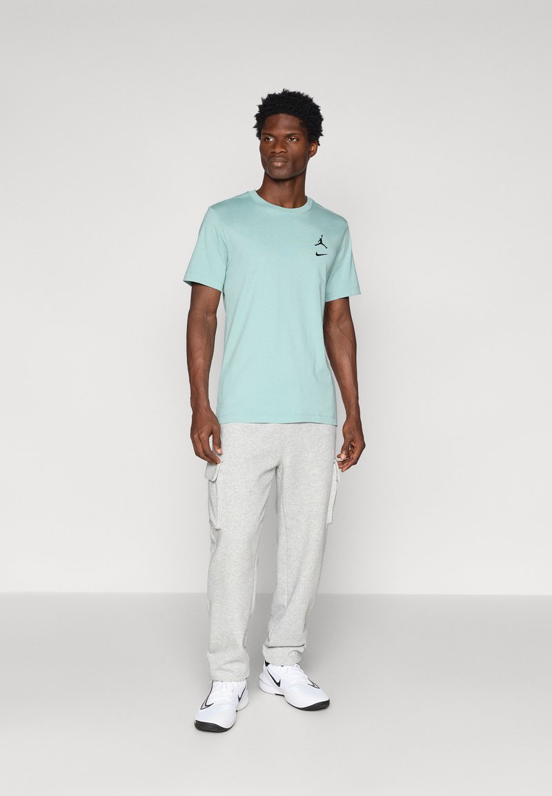 Camiseta de manga corta verde menta con logo negro, combinada con pantalones de chándal cargo grises claros; unas zapatillas deportivas blancas completan el atuendo.