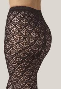 Närbild av en persons nedre torso och ben som bär svarta spetsstrumpbyxor med ett skaldjur- och fläktmönster mot en neutral bakgrund.