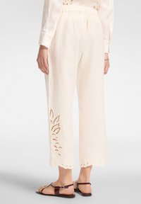 Pantaloni culotte bianchi in lino con dettagli a fiori ritagliati e orlo scallop. Presentano una vita elastica e una consistenza morbida. Abbinati a sandali marroni.