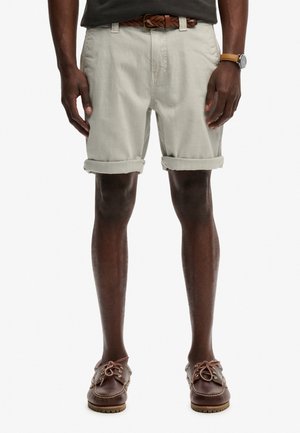 Mężczyzna ubrany w beżowe spodnie krótkie z podwiniętymi nogawkami, brązowy pleciony pasek, brązowy skórzany zegarek oraz brązowe skórzane buty żeglarskie na białym tle.