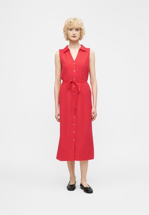 Γυναίκα με σγουρά ξανθά μαλλιά που φοράει ένα αμάνικο κόκκινο φόρεμα με κουμπιά και ζώνη στη μέση, καθώς και μαύρες επίπεδες γόβες, στέκεται μπροστά σε λευκό φόντο.