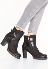 Botines de tacón alto negros con parte trasera texturizada, correas con hebilla y cremallera lateral, combinados con jeans ajustados de corte deshilachado en gris claro.