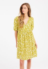 Kurzes, ärmelloses Kleid mit V-Ausschnitt aus leicht olivgrünem Stoff mit weißen botanischen Mustern. Ausgestellter Rock mit gerafftem Bund für eine lockere Passform.
