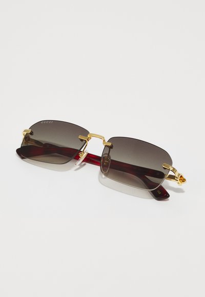 Gafas de sol Gucci con lentes de color marrón, acentos metálicos dorados y brazos transparentes rojos. Diseño rectangular elegante con el logotipo de la marca visible.