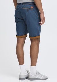 Blå shorts med rullede brune manchetter, to baglommer med knapper og en brun detalje i taljen. Båret med lysegrå sneakers og hvide sokker.