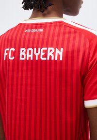 Άτομο που φορεί κόκκινη φανέλα της FC Bayern με κάθετες ρίγες και τη φράση «MIA SAN MIA» κοντά στο λαιμό.