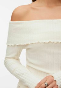 Krēma krāsas pieguļošs džemperis ar garām piedurknēm, ar viļņotu apakšu un teksturētu audumu. Rokā redzams sudraba gredzens.