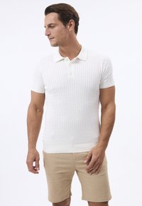 Camisa polo blanca de manga corta con un patrón de punto de cable texturizado y un collar suave. Combinada con pantalones cortos beiges, que muestran una silueta ajustada.