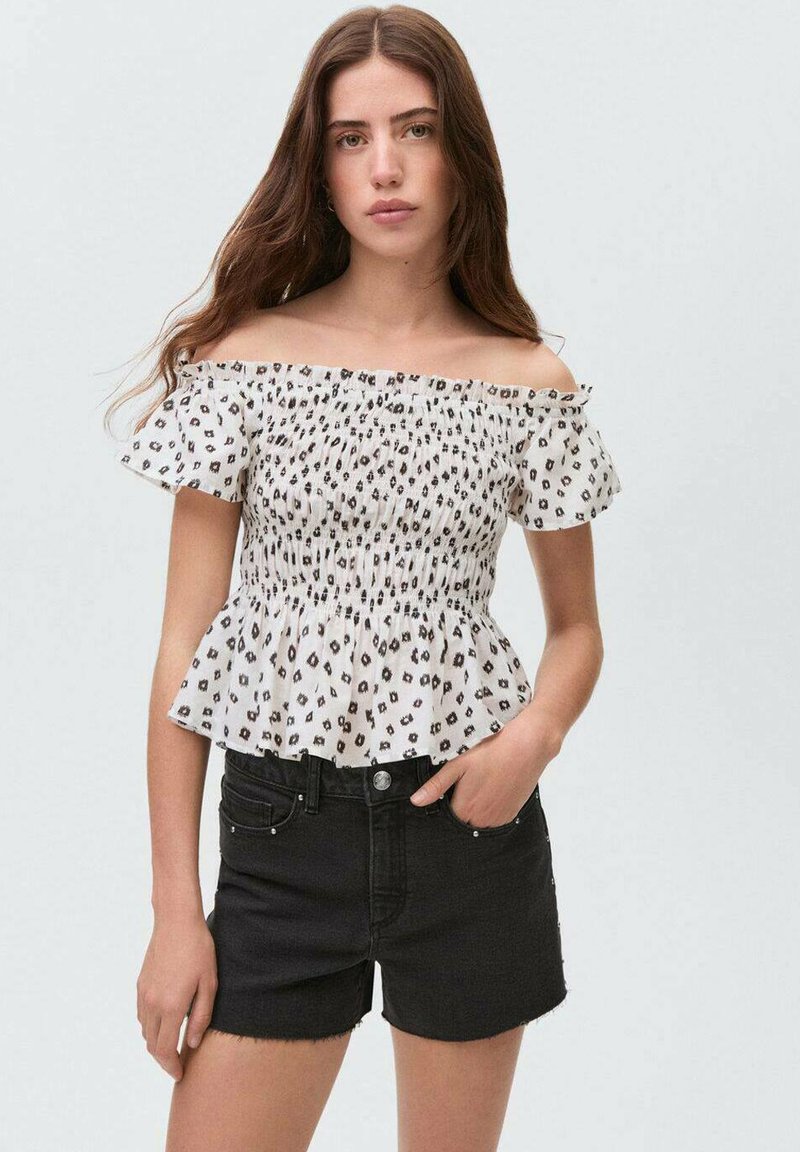Top s spuščenimi rameni in peplum krojem v beli barvi s črnimi cetami leoparda, ki vključuje nabranine in pufaste rokave, v kombinaciji z črnimi denin kratkimi hlačami.