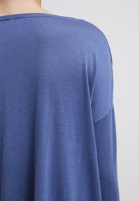 Pull en tricot bleu à coupe décontractée, présentant une texture lisse et un décolleté large. Le dos affiche un léger dégradé de couleur dans le tissu.