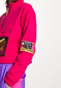 Persona vestida con una chaqueta de cremallera rosa brillante, con un bolsillo de malla negra y un parche en la manga que dice "Puma Helly Hansen Adaptado para el Deporte".