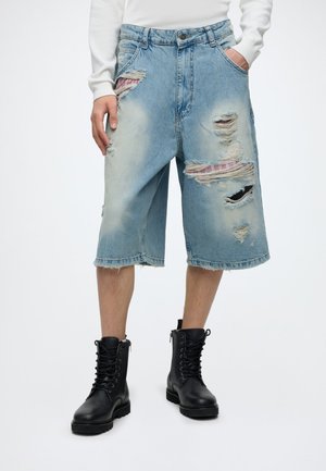 Shorts en jean bleu clair déchirés à hauteur du genou avec des patchs en tissu à carreaux rouges, portés avec des bottes noires à lacets et un haut blanc à manches longues.