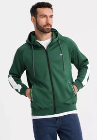 Felpa verde con zip e cappuccio, tasche frontali e accenti bianchi sui lati. Realizzata in un tessuto liscio e testurizzato. Coulisse sul cappuccio.