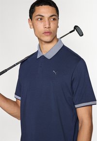 Μπλε κοντομάνικο πόλο με κολάρο και διακοσμητικές λεπτομέρειες στα μανίκια. Διαθέτει τριών κουμπιών μπροστινό άνοιγμα και κεντημένο λογότυπο στο στήθος.