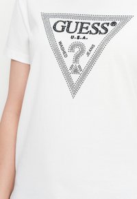 White t-shirt with a large triangle logo made of small studs, featuring "GUESS U.S.A.", a question mark, and the words "Washed Jeans".
