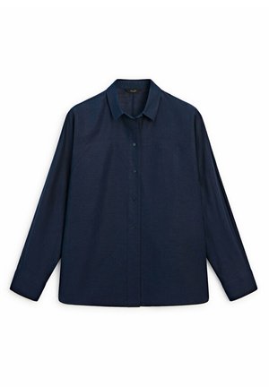 Person wearing a loose dark navy shirt, high-waisted wide-leg blue jeans, and black slip-on shoes, standing against a plain light wall.