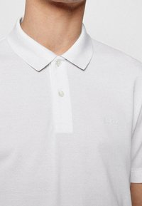 Polo blanc en tissu texturé, avec un col plat, deux boutons et un logo en relief subtil sur la poitrine gauche.