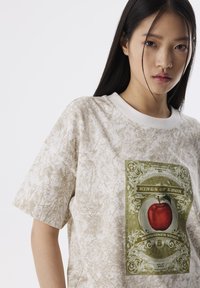 Jasno beżowy t-shirt o teksturowanym wykończeniu, z centralnym grafiką w zieleni i czerwieni przedstawiającą jabłko oraz ozdobnym napisem.