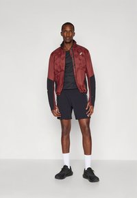 Bordowa sportowa kurtka z teksturowaną górą, czarne długie rękawy; zestawiona z czarnym T-shirtem i szortami. Czarne buty i białe skarpetki dopełniają wygląd.