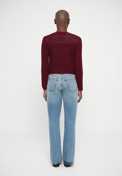 Sweater tricotado cor burgundy com um padrão texturizado, combinado com jeans claros de perna larga. A parte de trás apresenta detalhes de trama aberta nos ombros.