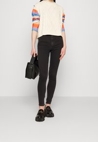 Suéter de punto trenzado en color crema con mangas de rayas coloridas, jeans skinny negros, bolso negro y mocasines negros gruesos con detalle de cadena dorada.