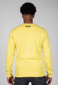 Sudadera amarilla con cuello redondo y mangas largas, con el logo "WRONG FRIENDS" impreso en la parte superior de la espalda. Textura suave.